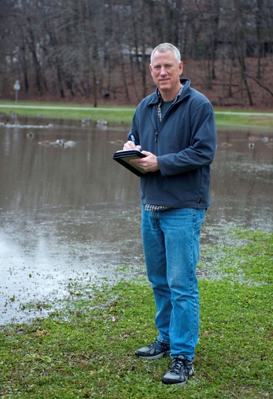 A man in blue jeans stands beside a pond, writing on a clipboard